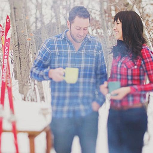 Two people in light layers enjoying hot drinks by a hire in the snow.